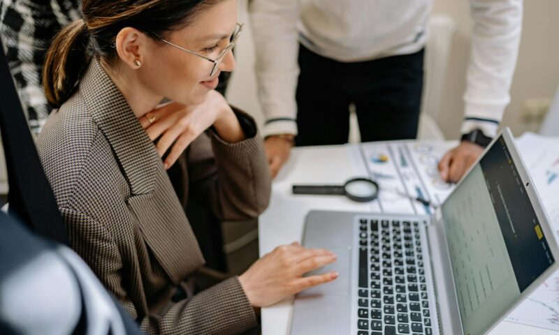 Businesswoman using laptop at office desk surrounded by colleagues, collaborating on project.